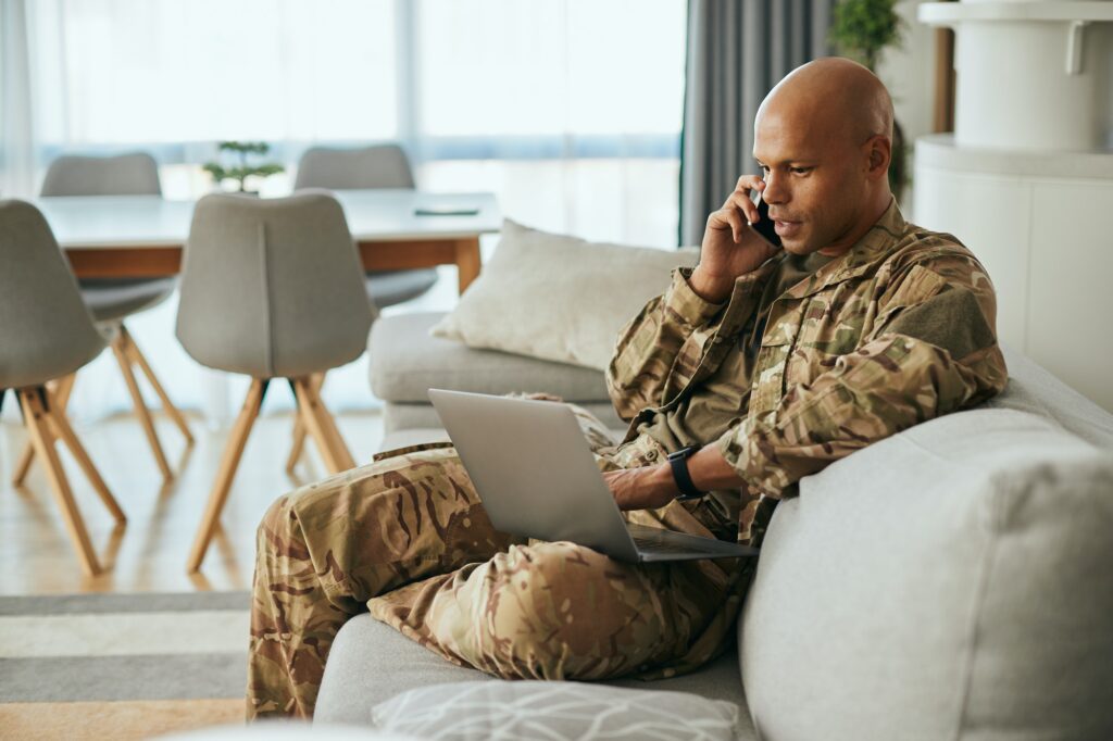 Black military man using laptop while talking on the phone at home.